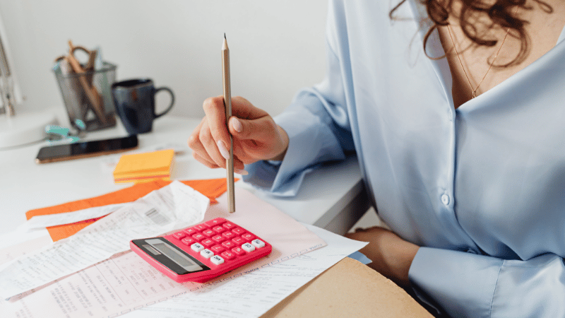 A businesswoman in a blue shirt sits at a desk covered with financial documents, holding a pencil and using a pink calculator.